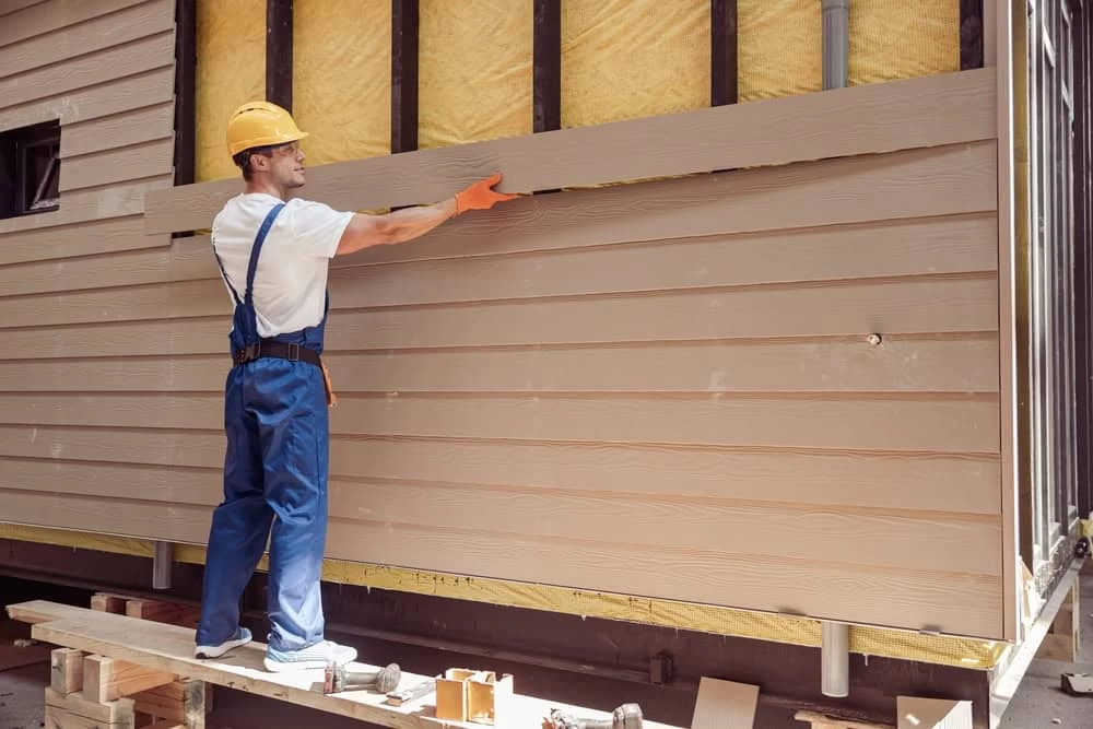 A construction worker in blue overalls and a yellow hard hat installs siding on a building.