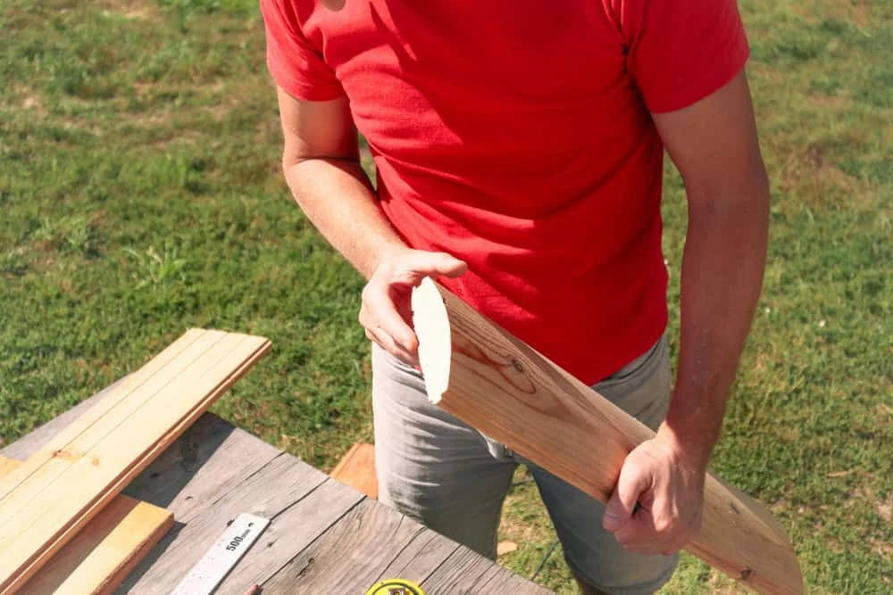 A person in a red shirt and gray shorts is sanding a piece of wood at an outdoor workbench. The table has additional wooden planks and a measuring tape. The background is grass.