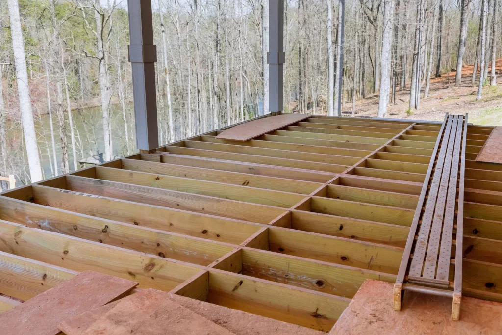 A wooden deck under construction with exposed beams and joists is set in a wooded area by a lake. Sheets of plywood are scattered around, and metal scaffolding planks rest on top. The background features tall trees and a glimpse of water.