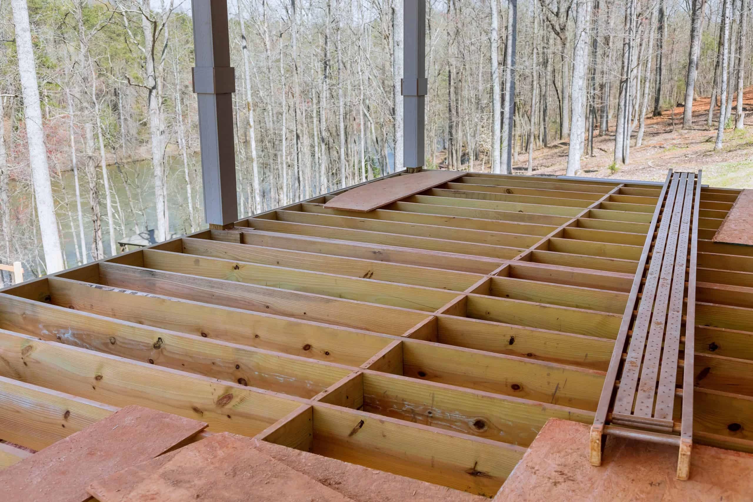 A wooden deck under construction with exposed beams and joists is set in a wooded area by a lake. Sheets of plywood are scattered around, and metal scaffolding planks rest on top. The background features tall trees and a glimpse of water.