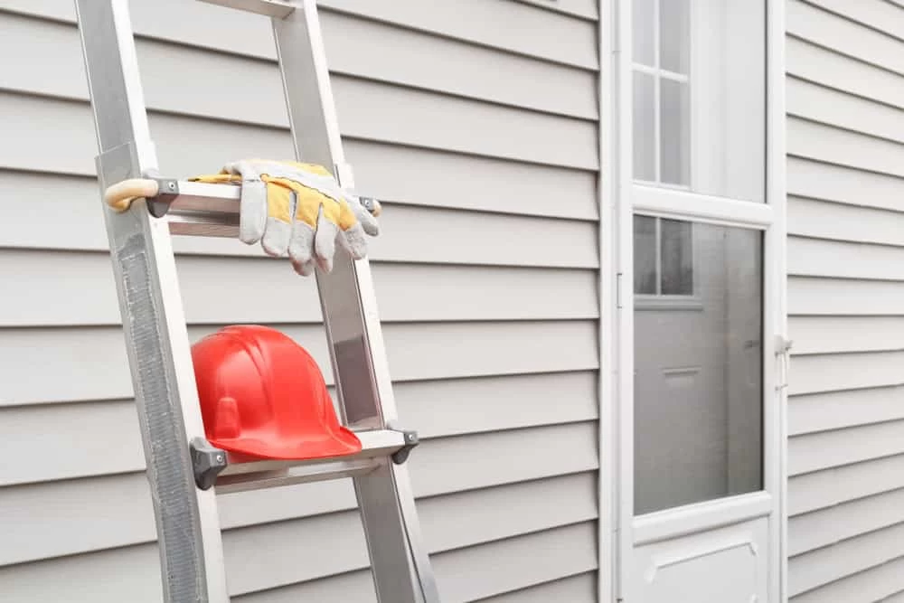 A ladder is leaning against a beige vinyl-sided house near a white door. A pair of work gloves and a red hard hat rest on the ladder.