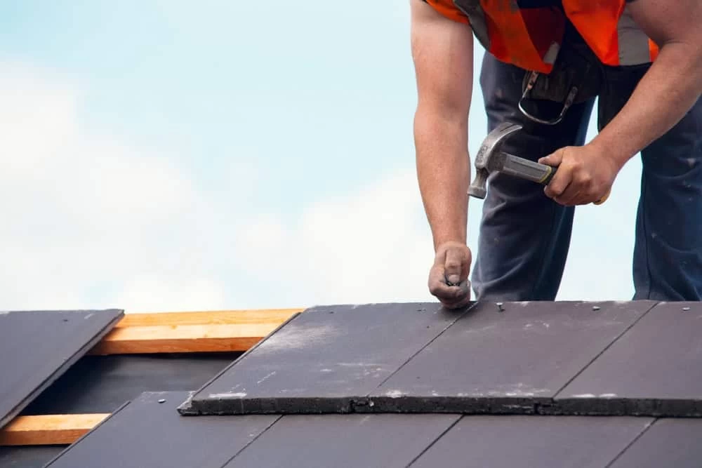 A construction worker wearing an orange safety vest is using a hammer to nail down dark roofing tiles on a roof. The sky is clear in the background, and wooden beams are visible under the tiles.