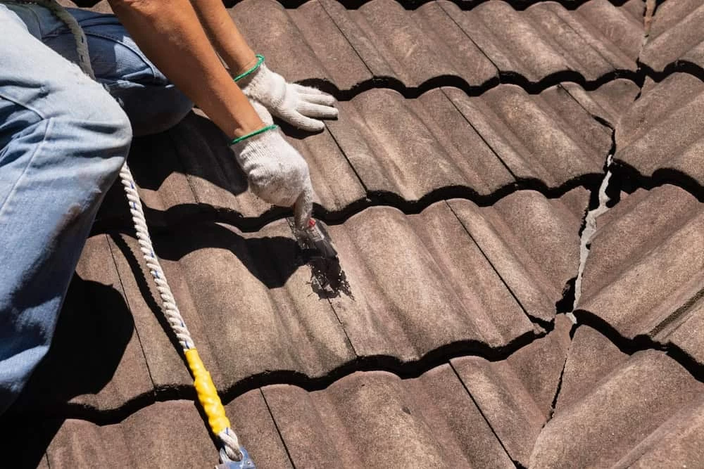 A person wearing gloves is applying cement to repair cracks on a brown tiled roof. They are seated, with safety gear visible, including a rope. The focus is on their hands and the damaged roof tiles.