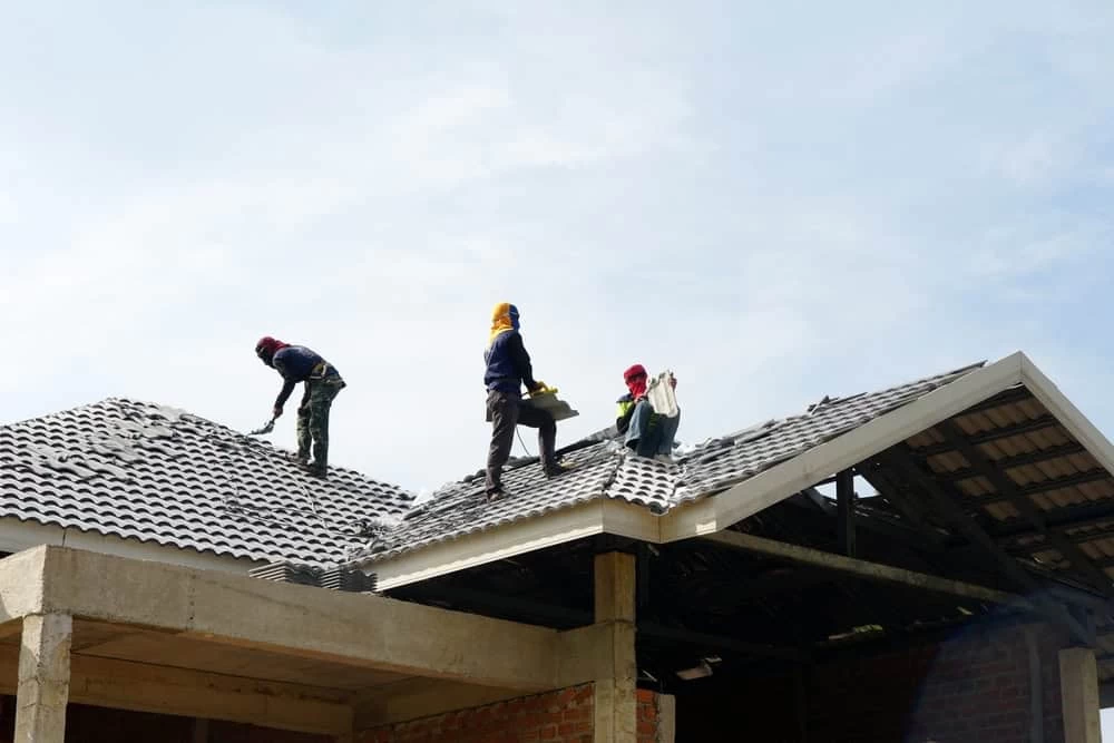 Three construction workers are on top of a tile roof, working on its installation. They're wearing protective clothing and hats, with a clear blue sky in the background.