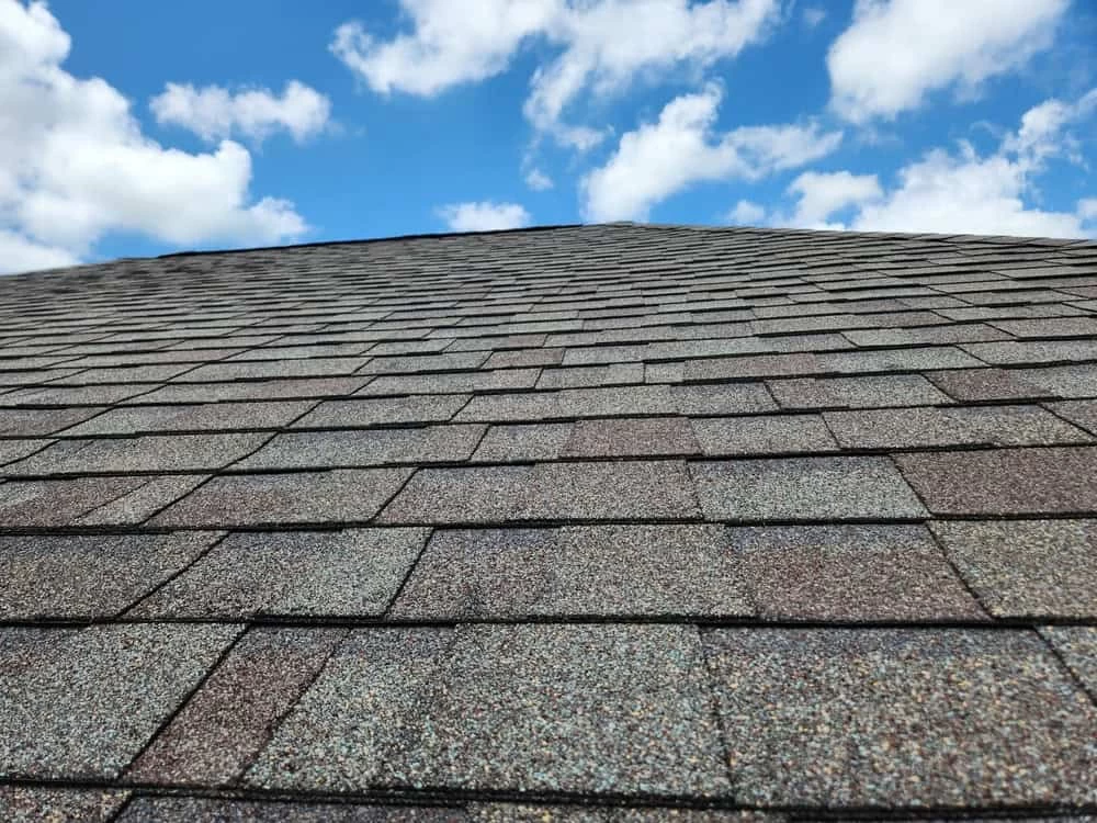 Rooftop with gray shingles extending towards a blue sky filled with scattered white clouds.