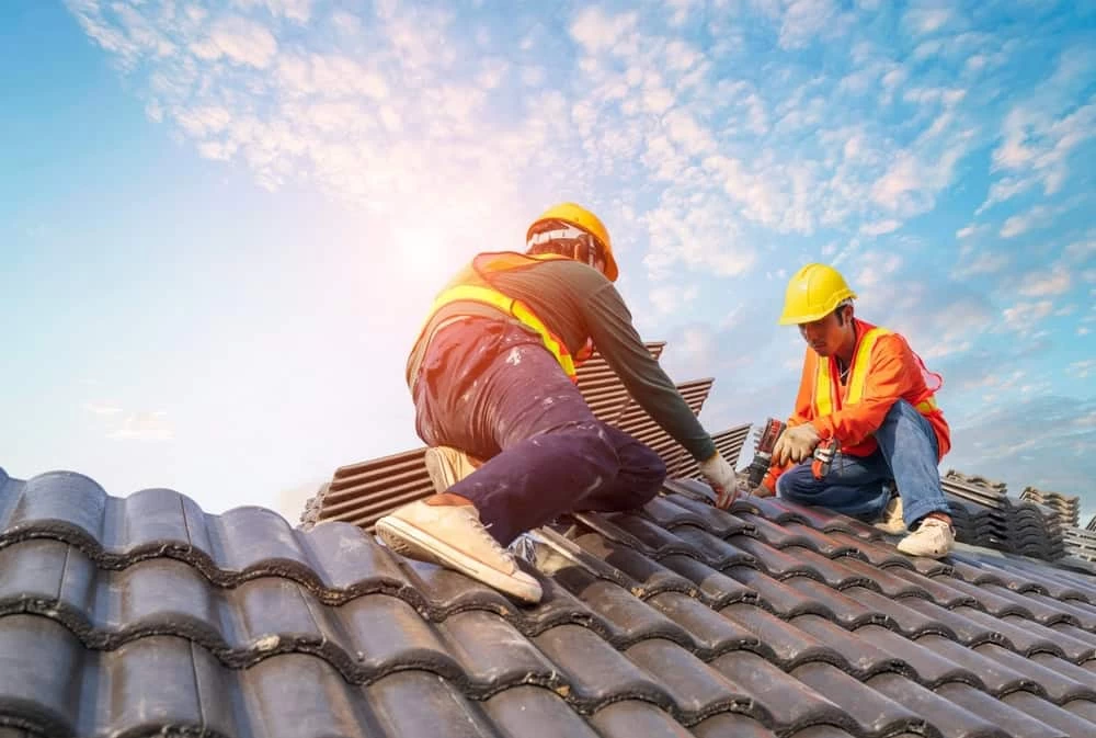 Two construction workers wearing safety gear are installing dark gray tiles on a sloped roof under a blue sky with scattered clouds. One worker is handling tiles, while the other is securing them, both in orange vests and yellow helmets.