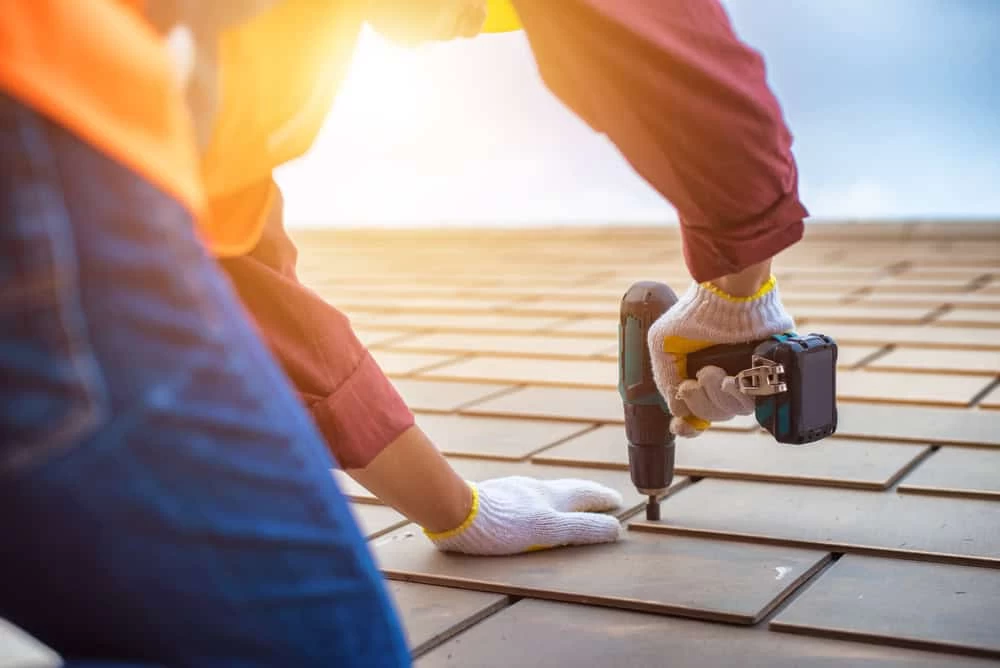 A person wearing gloves and a long-sleeved shirt uses a power drill to secure a tile on a roof. The sun shines brightly in the background, highlighting the construction work in progress.