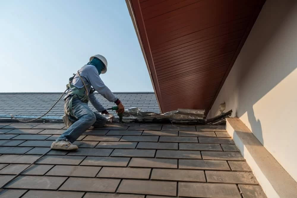A construction worker wearing safety gear is repairing a roof. They're adjusting shingles near the edge of the roof beneath a sloped overhang. The worker is tethered with a safety harness, and the sky is clear.