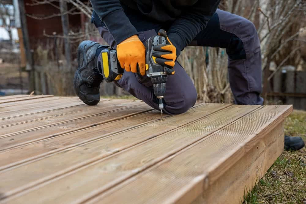 A person wearing gloves and work clothes kneels on a wooden deck, using a power drill to install or secure a screw into one of the deck boards outdoors.