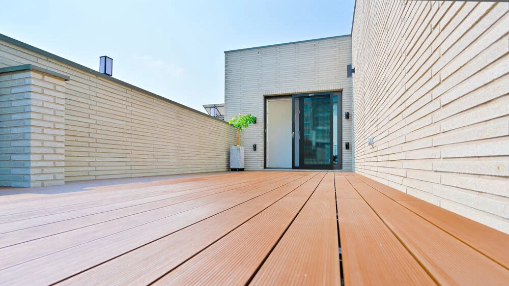 A modern rooftop terrace with brown wooden flooring, light-colored brick walls, a glass door, and a small potted plant, all under a clear blue sky.