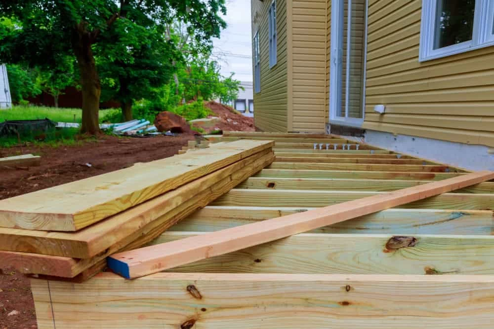 A partially built wooden deck is attached to a tan-sided house, with boards and beams placed across the frame. Piles of lumber rest on top, and a tree and soil are visible in the yard.